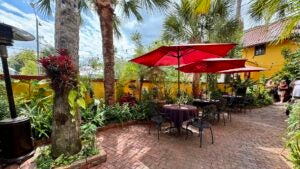 Outdoor space with yellow stone walls and a cobblestone floor with black patio furniture and red umbrellas surrounded by plants